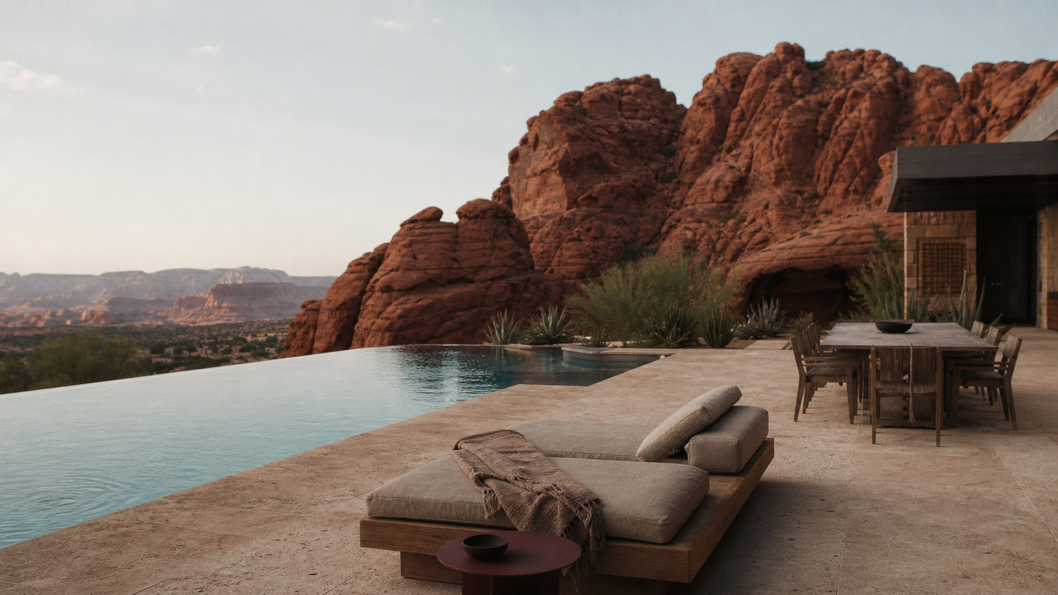 Desert estate with an infinity pool, lounge chairs and a dining table beneath red rock formations.