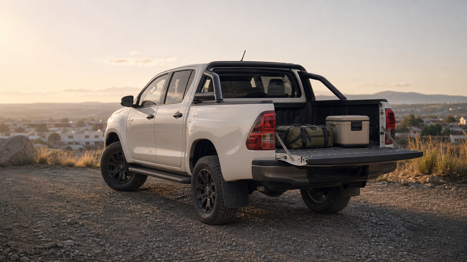 A Toyota Hilux Revo 4×4 pickup parked on a gravel road, with a duffel bag and cool box in its load bed.
