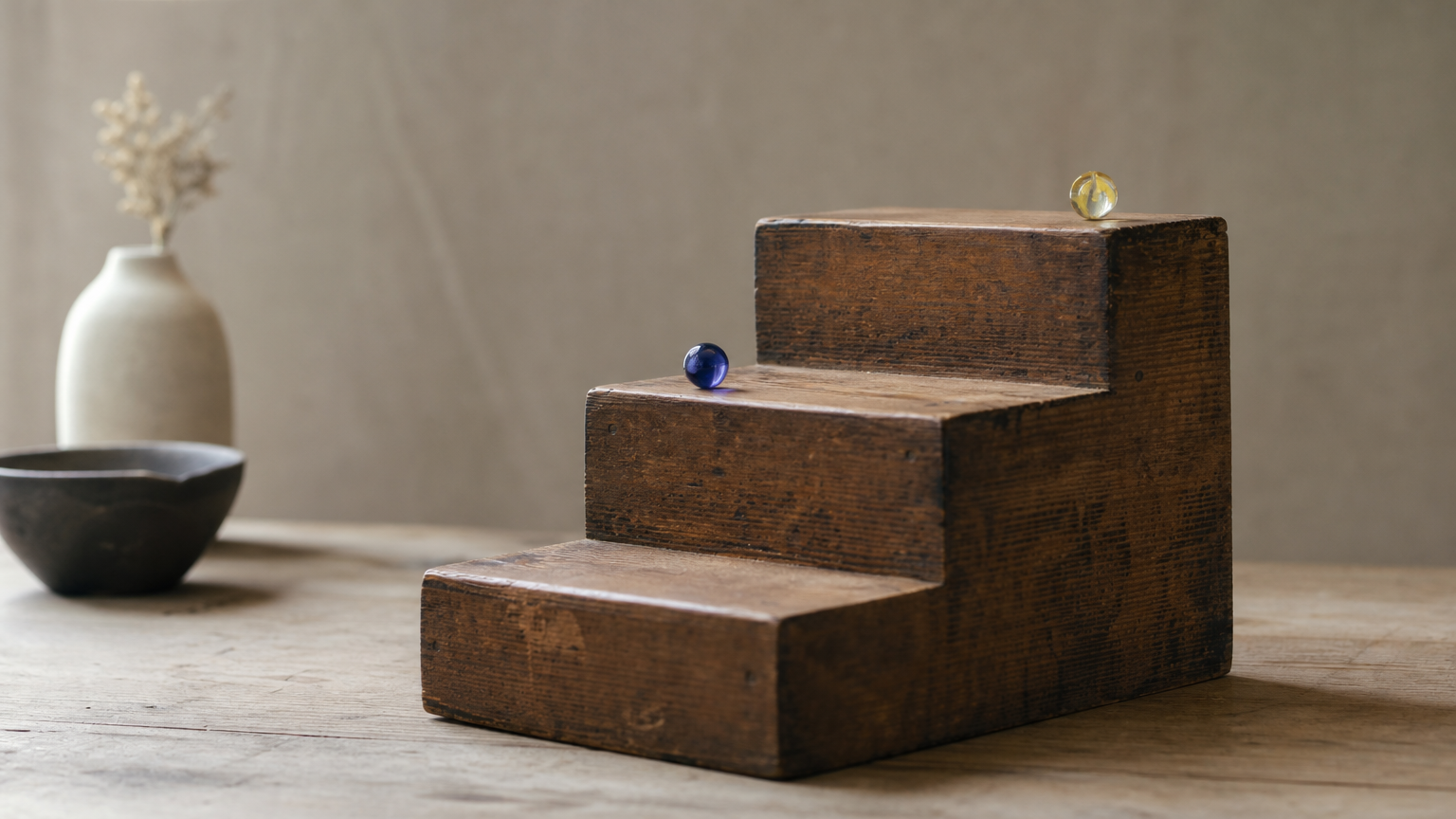A three-step wooden staircase from the side, with one marble on the top step and one on the middle step.